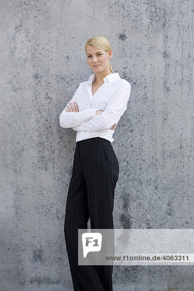 Junge blonde Frau im Business-Look steht mit verschränkten Armen vor einer grauen Wand