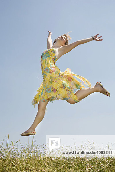 Junge langhaarige Frau springt im gelben Kleid vor blauem Himmel