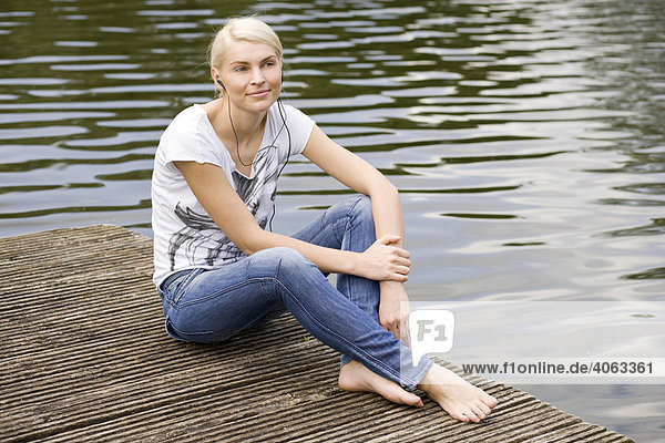 Smiling young blonde woman sitting on a wharf at a lake and listening to music