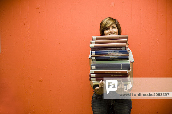 Junge dunkelhaarige Frau  Studentin  steht mit einem Stapel Bücher in den Händen vor einer orange-roten Wand