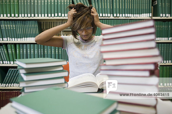 Junge dunkelhaarige Frau  Studentin  sitzt verzweifelt mit vielen Büchern an einem Tisch vor einem Bücherregal in einer Bilbiothek