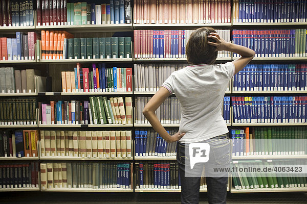 Junge dunkelhaarige Frau  Studentin  steht ratlos vor einem Bücherregal in einer Bibliothek