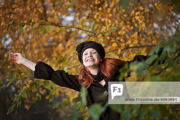 Young red haired woman wearing a woolen hat and scarf relishing autumn with outstretched arms
