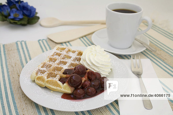 Waffle topped with powdered sugar  cherries and whipped cream served with a cup of coffee