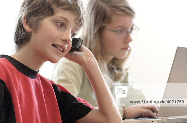 Ein Junge 10 Jahre alt telefoniert mit einem Handy und seine Schwester 13 Jahre alt sitzt an einem Laptop