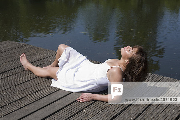 Frau im weißem Kleid liegt auf einem Holzsteg und genießt die Sonne