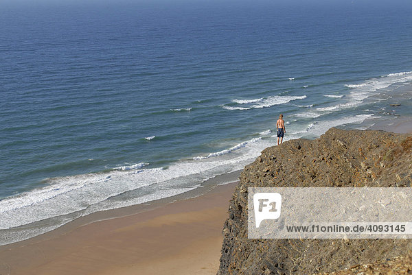 Junger Mann steht auf einem Felsen hoch über dem Meer und schaut auf den Ozean  Atlantik  Algarve  Portugal