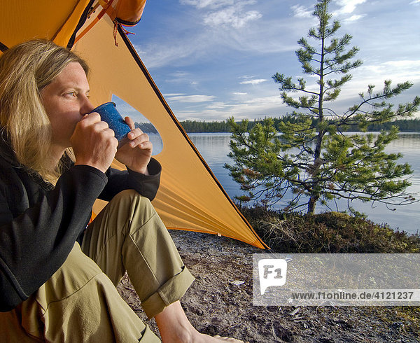 Frau sitzt vor einem Zelt  blickt in die Ferne und trinkt aus einer Tasse  Femundsmarka Nationalpark  Femundsmark  Norwegen