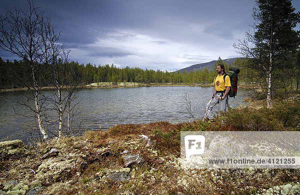 Man wearing backpack standing in front of a lake under cloudy sky  Jotunheimen National Park  Vaga  Oppland  Norwegen