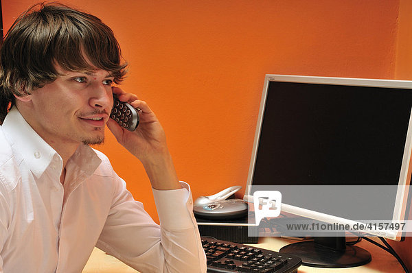 Junger Mann sitzt am Schreibtisch mit Computer und telefoniert