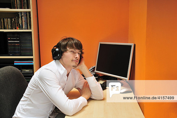 Young man wearing headphones sitting at desk in front of computer  listening to music  contemplating