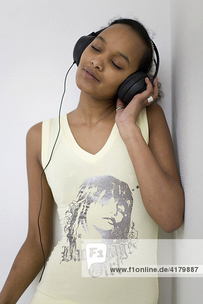 Junge dunkelhäutige Frau lehnt entspannt an einer Wand und hört über große Kopfhörer Musik