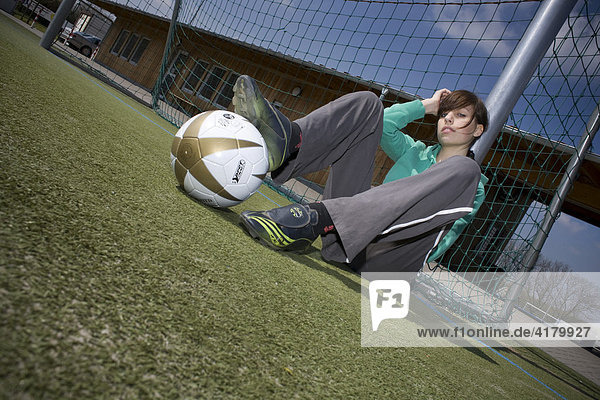 Junge Frau sitzt mit einem Fußball an einem Torpfosten gelehnt