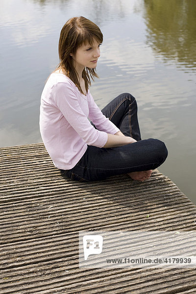 Junge Frau sitzt auf einem Holzsteg am Wasser und blickt auf das Wasser