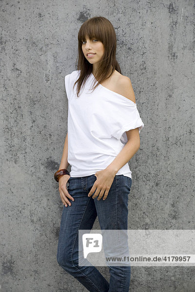 Junge dunkelhaarige Frau steht mit Jeans und weißem Top vor einer Wand und blickt entspannt in die Kamera