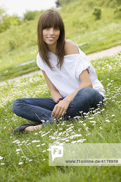 Junge dunkelhaarige Frau mit Jeans und weißem Top sitzt auf einer sommerlichen Wiese und blickt freundlich in die Kamera