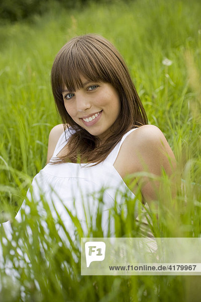 Junge dunkelhaarige Frau in einem weißen Kleid liegt entspannt auf einer Wiese  genießt den Sommer  blickt lächelnd in die Kamera