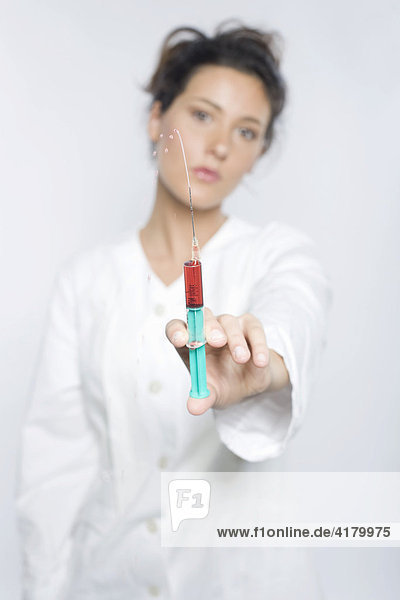 Junge Frau mit weißem Laborkittel hält eine Spritze mit roter Flüssigkeit in die Kamera