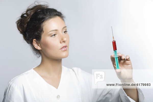 Junge Frau mit weißem Laborkittel hält eine Spritze mit roter Flüssigkeit in den Händen und blickt konzentriert