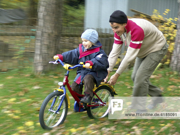 Erste Fahrversuche der vierjaehrigen Mädchen auf dem neuen Fahrrad ohne Stuetzraeder mit Hilfe des Vaters im Garten.
