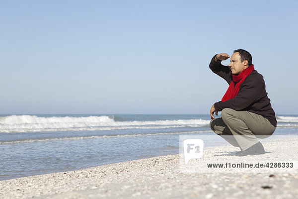 Mann hockt am Strand und schaut auf den Ozean.