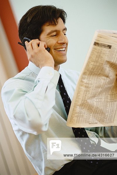 Businessman using phone and holding up financial section of newspaper  smiling