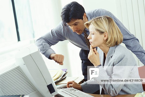 Businessman leaning over female colleague's shoulder looking at monitor