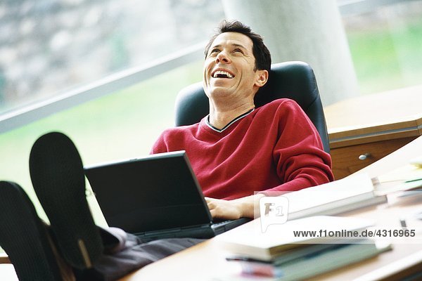 Mann sitzt am Schreibtisch mit erhobenen Füßen  hält Laptop auf dem Schoß  lacht