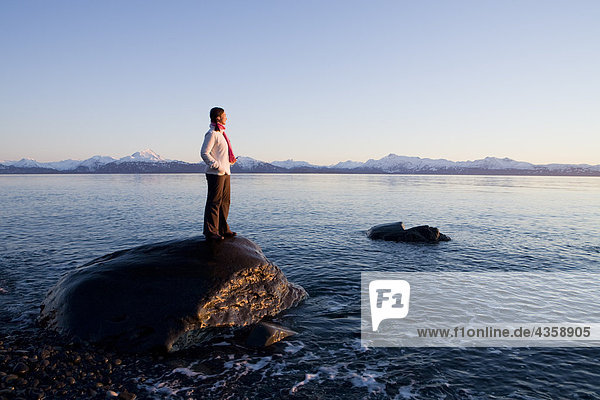 Woman stands on rock at Bishops Beach viewing Kachemak Bay in Homer  Kenai Peninsula  Southcentral Alaska  Winter