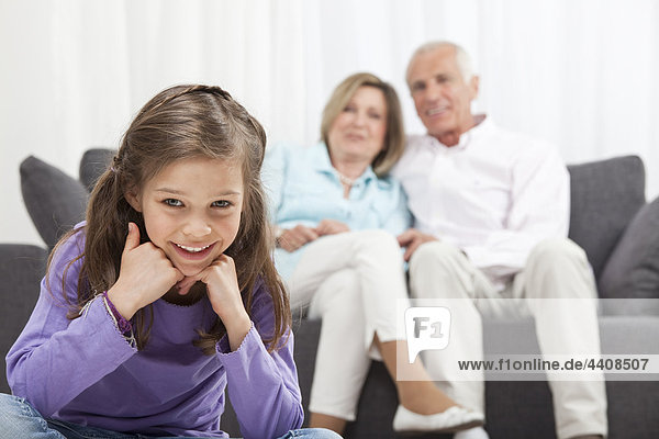 Enkelin (6-7) lächelnd mit Kopf in der Hand und Großeltern im Hintergrund