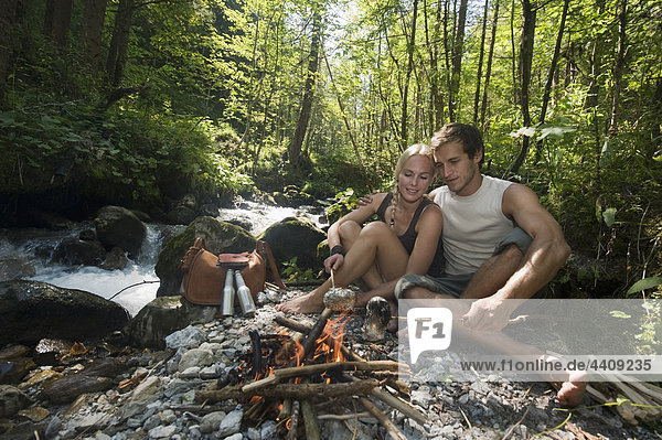 Austria  Steiermark  Young couple sitting at camp fire by stream in forest