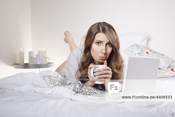 Frau liegt auf dem Bett und hält Kaffeetasse mit Laptop  Stirnrunzeln