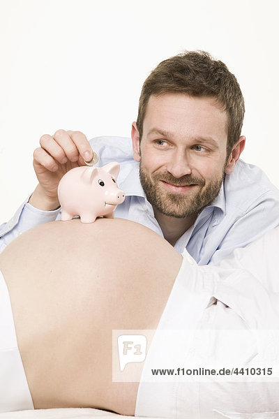 Mann  der Geld in Sparschwein auf den Bauch einer schwangeren Frau legt  Nahaufnahme