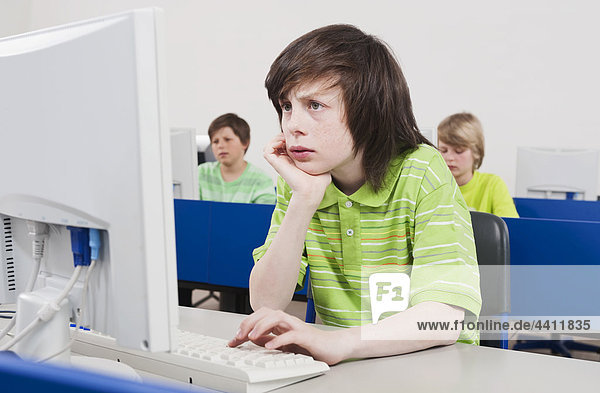 Boy (12-13) using computer with hand on chin and students in background