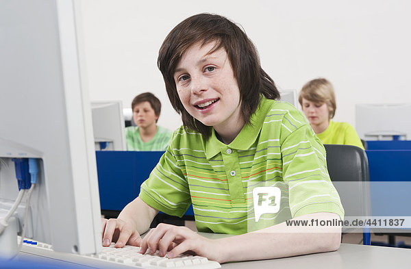 Boy (12-13) smiling and using computer with students in background