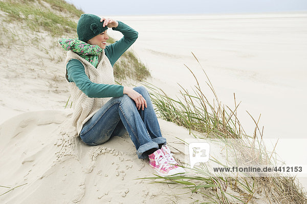 Deutschland  St. Peter-Ording  Nordsee  Frau auf Sanddünen sitzend und wegschauend  lächelnd