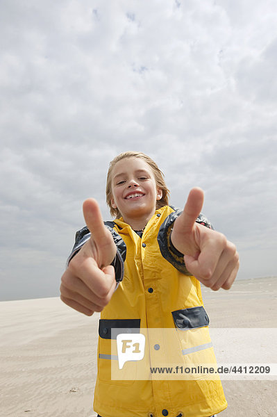 Nordsee  Junge (8-9) im Regenmantel mit Daumen nach oben Schild am Strand