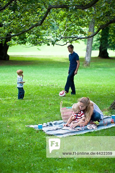 Mother reading book to daughter and father playing soccer with son in the park