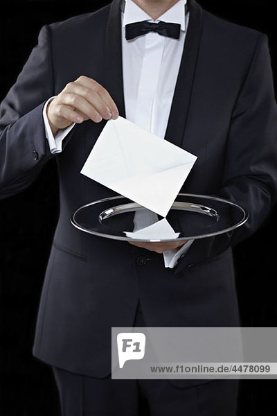 Mittelteil eines Mannes  der einen Smoking trägt und einen Umschlag über einer Silberplatte hält.