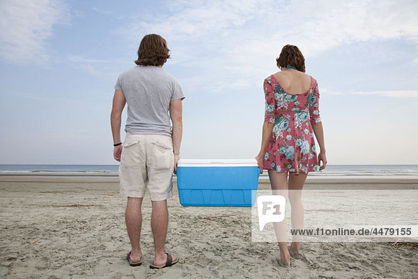 Ein junges Paar trägt eine Kühlbox zusammen am Strand.