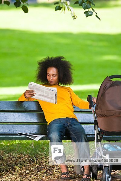 Junger Mann mit Kinderwagen auf Bank sitzen und lesen Zeitung