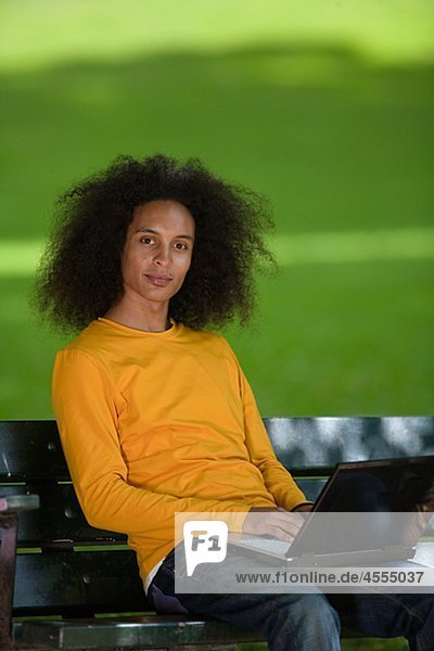 Smiling young man with afro hair sitting on bench with laptop