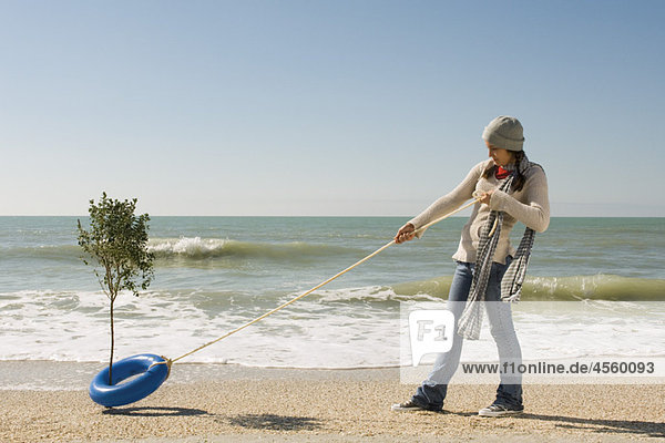 Preteen Mädchen ziehen Seil am Rettungsring um den Baum  der am Rand des Wassers am Strand wächst.