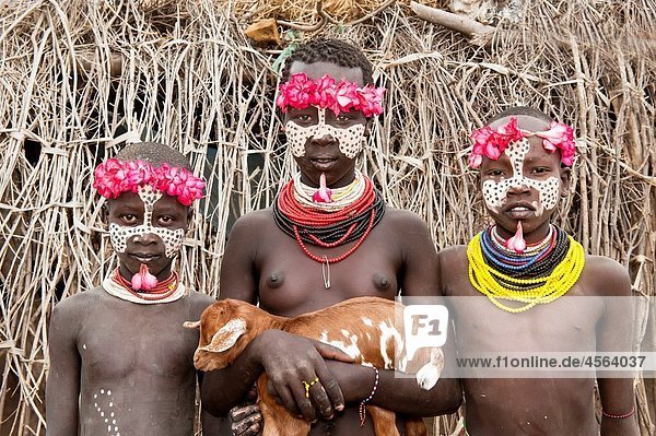 Three Karo girls with a floral headband  facial paintings  colorful necklaces  lip piercing and a goat in the arms  Omo river valley  Southern Ethiopia