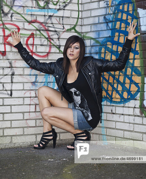 Junge Frau mit dunklen Haaren  Hotpants  schwarzer Lederjacke und hochhackigen Schuhen posiert vor Graffitiwand