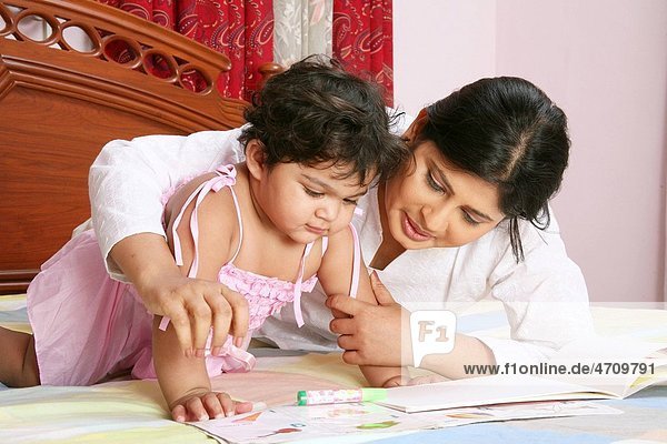 young mother telling story and teaching to baby girl of one and half year lying on bed with book MR 687 A and 687 B