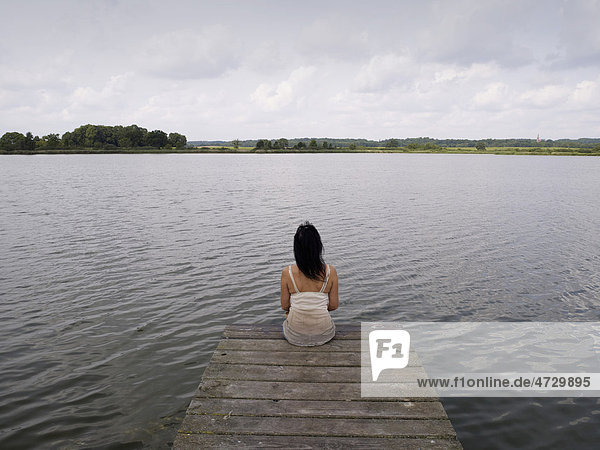 Frau sitzt auf einem Holzsteg an einem See der Mecklenburgischen Seenplatte  Mecklenburg-Vorpommern  Deutschland  Europa