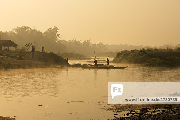 Einbaum mit stakendem Fährmann überquert kleinen Fluss in Morgenstimmung mit aufgehender Sonne und Nebel  Chitwan Nationalpark  Nepal  Asien