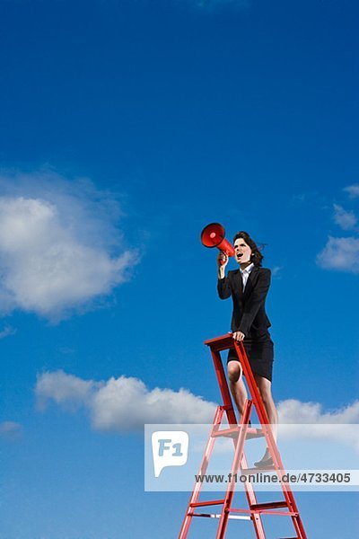 Businesswoman standing on top of red ladder against blue sky and shouting through red megaphone