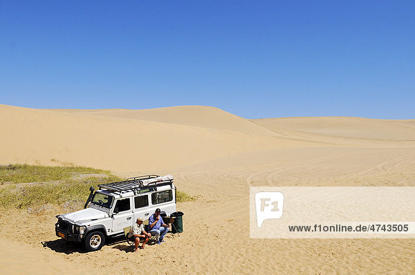 Two men sitting in front of a Landrover Defender off-road vehicle in the dunes of the Namib Naukluft National Park  part of the Namibian Skeleton Coast National Park  Skeleton Coast  Namib Desert  Namibia  Africa